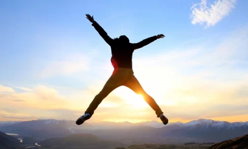 山の頂上で朝日を浴びながら両手を広げジャンプする男性のシルエット写真